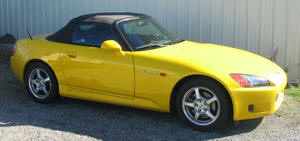 A yellow Honda S2000 roadster parked on gravel in front of a metal-sided building